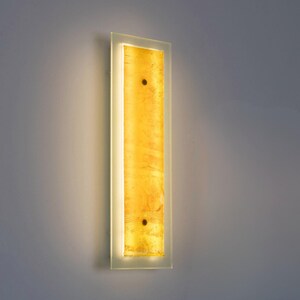 May include: A rectangular wall sconce featuring a central vertical panel of textured amber-colored glass framed by a clear glass border. The fixture emits a warm glow and is secured to the wall with two visible dark circular fasteners on the front.