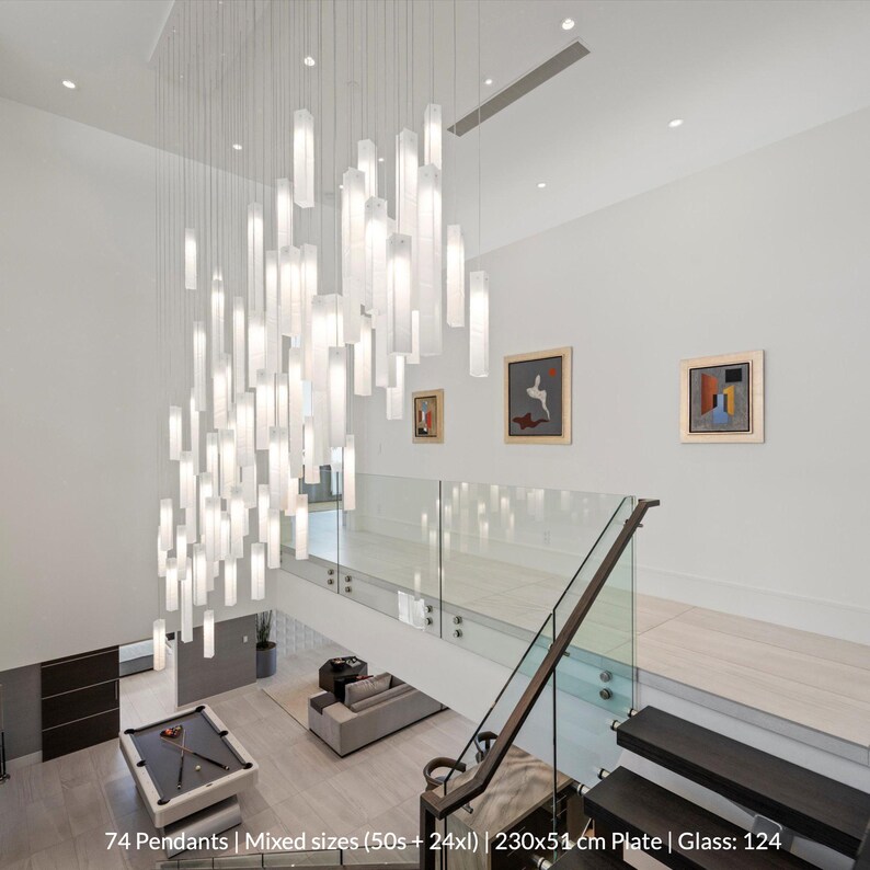 a modern and stylish living room with a pool table, glass railings, and a large hanging light fixture. The room has a minimalist design with white walls and a glass floor. There are also several paintings on the walls, adding an artistic touch to the space. The room appears to be well-lit and spacious, with a focus on the pool table and other decorative elements.