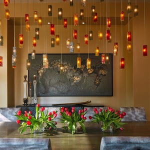 a modern dining room with a wooden table, gray upholstered chairs, and a large wall art piece. The table is set with red tulips in vases, and the room is illuminated by hanging lights.