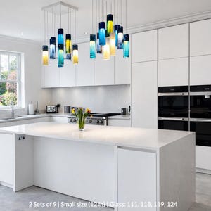 A modern, minimalist kitchen with white cabinets, a large island, and a hanging pendant light with colorful glass bottles.