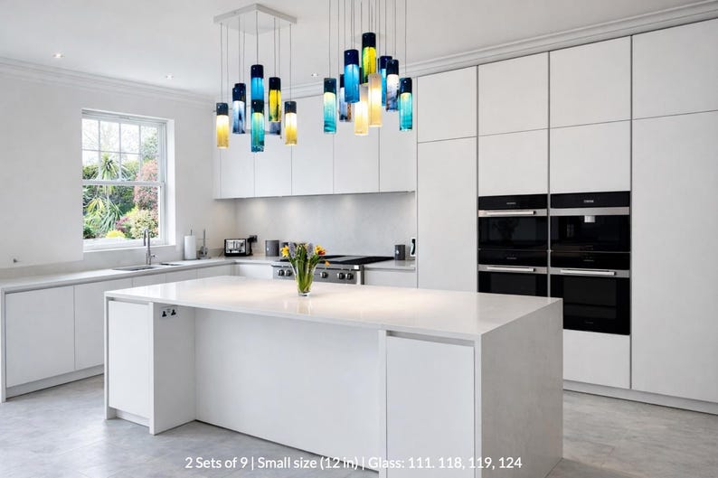 A modern, minimalist kitchen with white cabinets, a large island, and a hanging pendant light with colorful glass bottles.