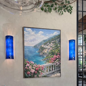 May include: A framed landscape painting of a coastal town with a blue sea and sky, flanked by two blue wall sconces. The painting features a white balustrade and pink flowers in the foreground. The sconces are cylindrical with a textured surface.