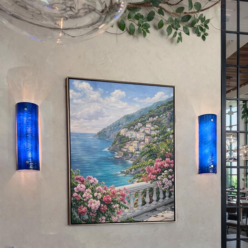 May include: A framed landscape painting of a coastal town with a blue sea and sky, flanked by two blue wall sconces. The painting features a white balustrade and pink flowers in the foreground. The sconces are cylindrical with a textured surface.