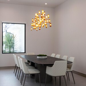 a modern dining room with a large rectangular table surrounded by white chairs. The table is set with a tray of greenery, and a hanging light fixture with numerous small lights illuminates the space. The room has a clean, minimalist design with white walls and a gray floor.
