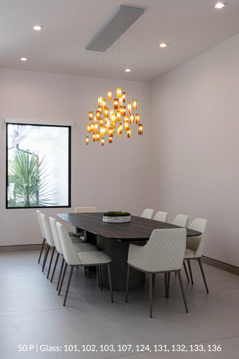 a modern dining room with a large rectangular table surrounded by white chairs. The table is set with a tray of greenery, and a hanging light fixture with numerous small lights illuminates the space. The room has a clean, minimalist design with white walls and a gray floor.