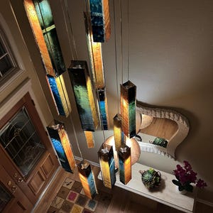 a room with a unique and colorful chandelier hanging from the ceiling, composed of multiple rectangular panels in various colors and shapes. The chandelier is suspended from the ceiling by metal rods, creating a striking visual effect. The room also includes a table with a vase of flowers, a mirror, and a potted plant, adding to the overall aesthetic of the space.