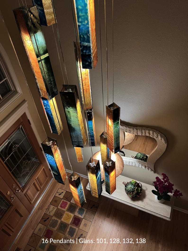 a room with a unique and colorful chandelier hanging from the ceiling, composed of multiple rectangular panels in various colors and shapes. The chandelier is suspended from the ceiling by metal rods, creating a striking visual effect. The room also includes a table with a vase of flowers, a mirror, and a potted plant, adding to the overall aesthetic of the space.
