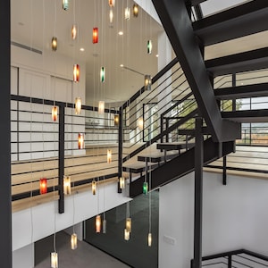 a modern interior design with a staircase and a hanging light fixture. The staircase has a black metal railing and is illuminated by a series of colorful lights suspended from the ceiling. The lights create a visually striking and contemporary atmosphere in the space.