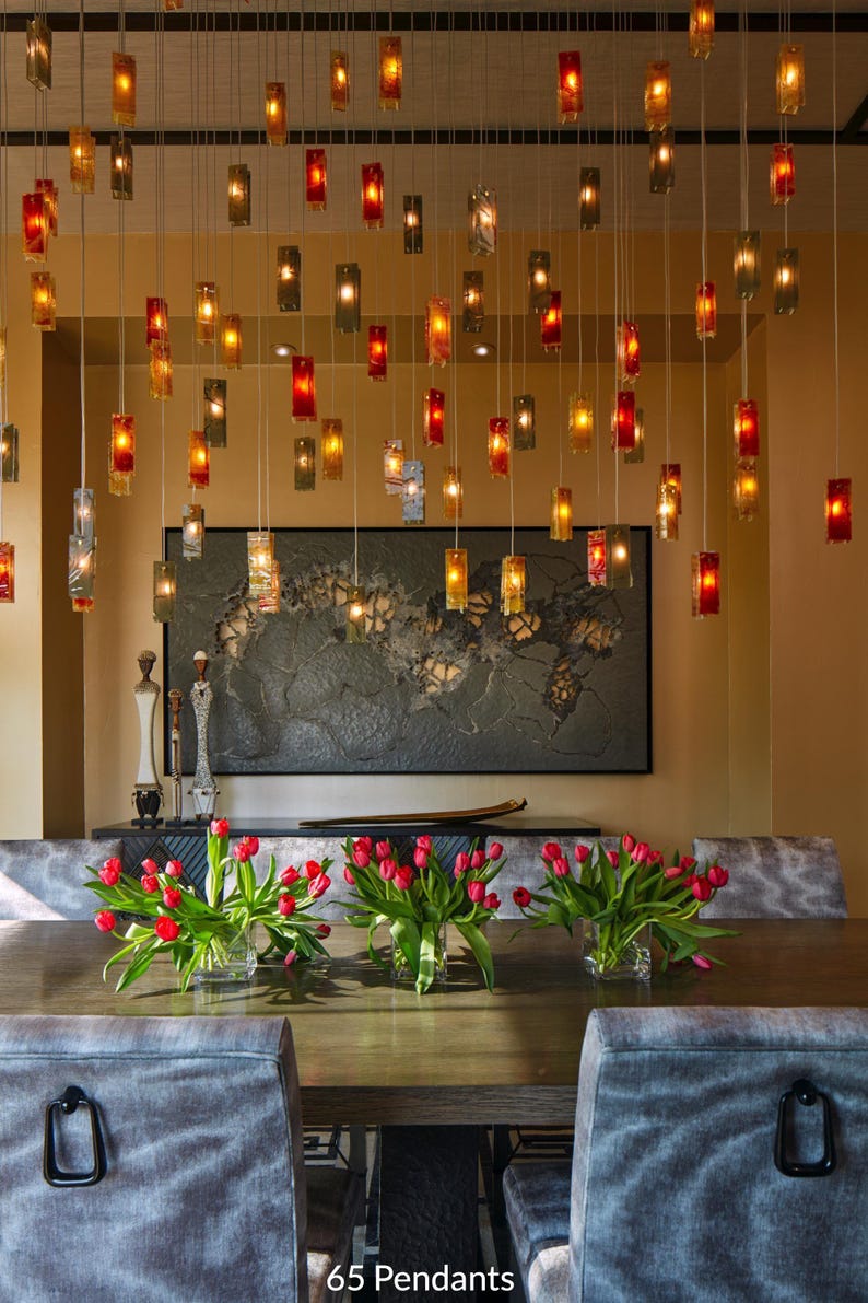 a modern dining room with a wooden table, gray upholstered chairs, and a large wall art piece. The table is set with red tulips in vases, and the room is illuminated by hanging lights.