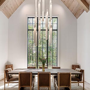 Puede incluir: Comedor moderno con una gran mesa rectangular de madera y seis sillas de cuero marrón. Una gran ventana ofrece vistas a un bosque. Una lámpara decorativa cuelga sobre la mesa. La habitación tiene una alfombra clara y un techo de madera.