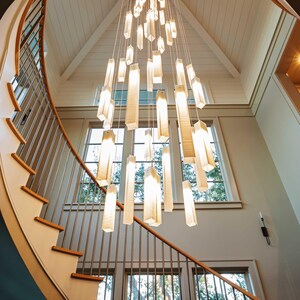 May include: A multi-tiered chandelier featuring numerous rectangular, translucent white glass light fixtures suspended at varying heights by thin wires, creating a cascading vertical arrangement within a high-ceilinged stairwell.