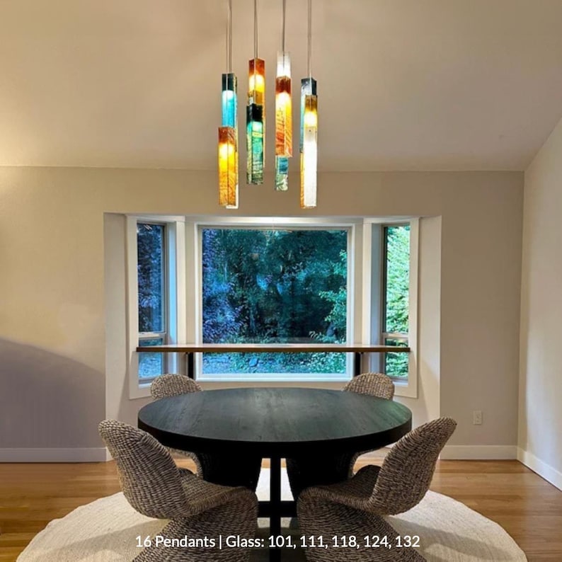 a dining room with a round wooden table and four chairs, all positioned in front of a large window that offers a view of trees. The room has a modern and minimalist design, with a chandelier hanging from the ceiling and a colorful pendant light fixture above the table.