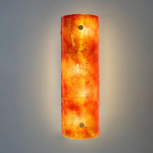 Puede incluir: Aplique de pared cilíndrico con un acabado texturizado en naranja y amarillo fuego. El aplique tiene dos pequeños acentos circulares oscuros y emite un brillo cálido. Diseñado para iluminación interior.