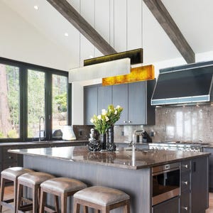 May include: A kitchen island with a dark brown stone worktop and dark wood cabinetry, featuring four stools with grey upholstered seats. Above the island hang three rectangular pendant lights in black, silver, and gold metallic finishes.