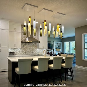 a modern kitchen with a large island and a set of white barstools. The kitchen has white cabinets and a backsplash with a geometric pattern. The island has a sink and a countertop with a sink and a coffee maker. The ceiling has hanging lights that are illuminated, creating a warm and inviting atmosphere.