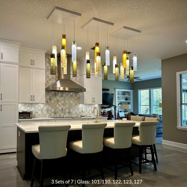a modern kitchen with a large island and a set of white barstools. The kitchen has white cabinets and a backsplash with a geometric pattern. The island has a sink and a countertop with a sink and a coffee maker. The ceiling has hanging lights that are illuminated, creating a warm and inviting atmosphere.