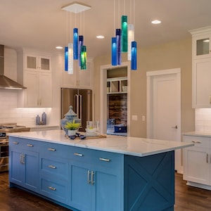 a modern kitchen with a large blue island and hanging glass pendant lights. The kitchen has white cabinets and a white tile backsplash, creating a clean and bright atmosphere.