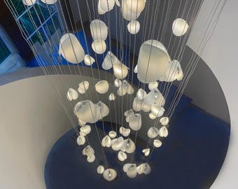 May include: A large chandelier featuring numerous white, petal-shaped glass light fixtures suspended at varying heights by thin wires. The translucent, organic-shaped shades are arranged in a cascading formation, creating a glowing, multi-layered lighting installation that hangs vertically above a dark blue spiral staircase.