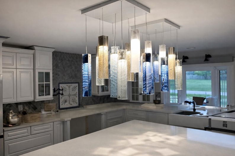 a modern kitchen with a large, stylish pendant light hanging from the ceiling. The light has a mix of blue and white glass elements, creating a visually appealing and contemporary design. The kitchen is well-equipped with white cabinets and a large island countertop, providing ample space for cooking and dining.