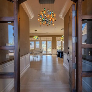 a spacious and well-lit entryway with a large wooden archway, a chandelier with colorful glass pieces hanging from the ceiling, and a piano visible in the background. The floor is made of wood, and the overall atmosphere appears to be elegant and inviting.