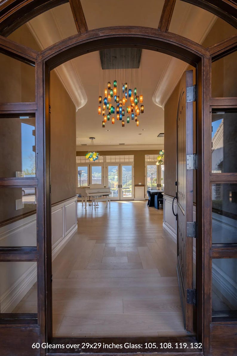 a spacious and well-lit entryway with a large wooden archway, a chandelier with colorful glass pieces hanging from the ceiling, and a piano visible in the background. The floor is made of wood, and the overall atmosphere appears to be elegant and inviting.