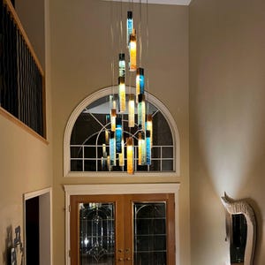 May include: A multi-pendant chandelier featuring numerous rectangular glass light fixtures suspended at varying heights. The glass pieces display a gradient of vibrant blue and warm amber tones, creating a vertical, cascading light installation.