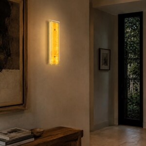 May include: A rectangular wall sconce with a warm, amber glow. The light fixture has a clear border and a textured, golden-yellow center. It is mounted on a neutral-colored wall, adding a touch of elegance to the space.