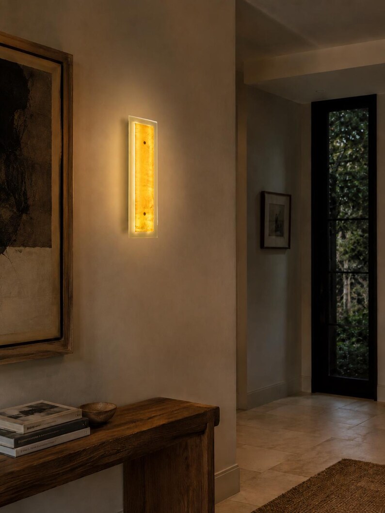 May include: A rectangular wall sconce with a warm, amber glow. The light fixture has a clear border and a textured, golden-yellow center. It is mounted on a neutral-colored wall, adding a touch of elegance to the space.