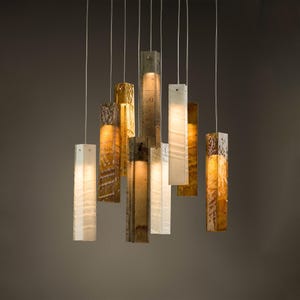 Puede incluir: Una lámpara de araña con múltiples colgantes que presenta varios accesorios de luz rectangulares y verticales suspendidos a diferentes alturas. Las pantallas están hechas de materiales texturizados y translúcidos en tonos ámbar, oro y blanco.