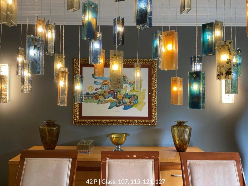 a dining room with a wooden table, three chairs, and a painting on the wall. The room has a modern and stylish design, with a mix of blue, green, and gold accents. The dining table is set with a bowl and a vase, and the chairs are arranged around the table. The painting on the wall adds an artistic touch to the space.