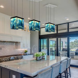 May include: Three square pendant lights hang from the ceiling, each featuring a vibrant blue and gold marbled glass shade. These fixtures are suspended by thin black cords above a kitchen island with a white marble countertop and gray chairs.