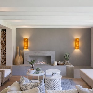 May include: A modern living room with a fireplace, two sconces, and two wicker chairs. The fireplace is built into a gray wall, and the sconces are lit. There are two small white tables and a long bench.