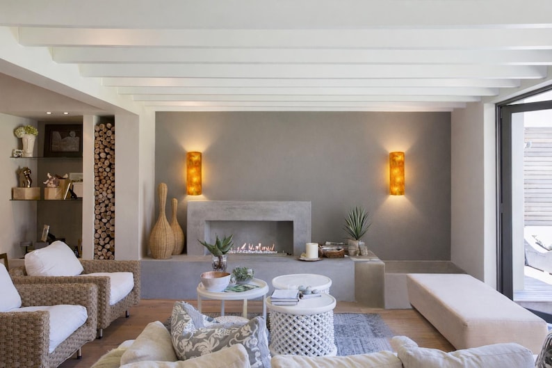 May include: A modern living room with a fireplace, two sconces, and two wicker chairs. The fireplace is built into a gray wall, and the sconces are lit. There are two small white tables and a long bench.