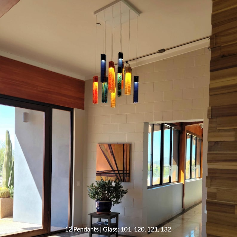 a modern interior with a hanging pendant light featuring colorful glass tubes suspended from the ceiling. The room has a wooden floor and white walls, and there is a painting on the wall. The pendant light is the focal point of the space, and the overall design appears to be contemporary and stylish.