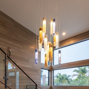 a modern interior design with a large, hanging chandelier made of glass and metal, featuring multiple hanging lights in various colors. The chandelier is suspended from the ceiling by strings, creating a striking focal point in the space. The room has a wooden floor and walls, and a staircase can be seen in the background.