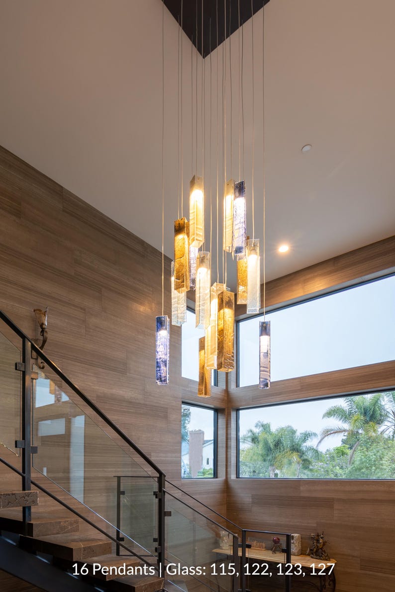 a modern interior design with a large, hanging chandelier made of glass and metal, featuring multiple hanging lights in various colors. The chandelier is suspended from the ceiling by strings, creating a striking focal point in the space. The room has a wooden floor and walls, and a staircase can be seen in the background.