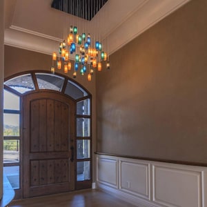 a large wooden door with a glass panel, and above it is a chandelier with colorful glass shades. The room has a beige wall and a wooden floor.