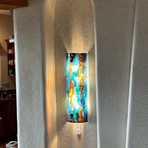 May include: A cylindrical wall sconce with a vibrant, abstract design in shades of blue, orange, and yellow. The light fixture is mounted inside a recessed alcove with a textured, off-white finish. A small shelf is below the sconce.