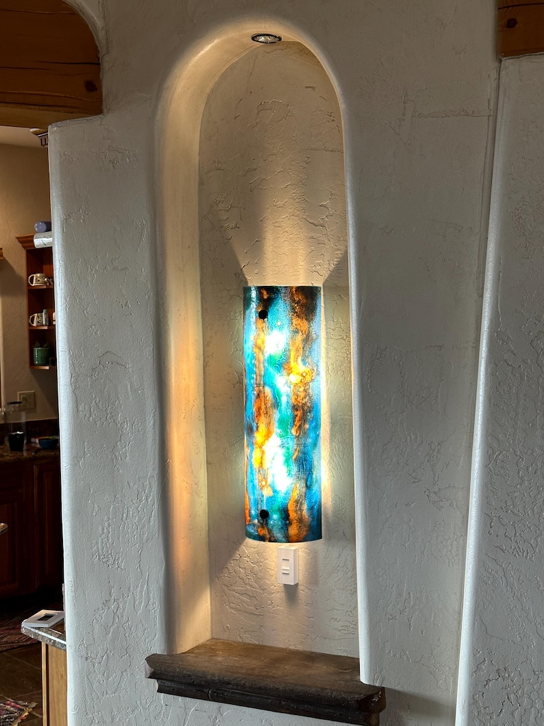 May include: A cylindrical wall sconce with a vibrant, abstract design in shades of blue, orange, and yellow. The light fixture is mounted inside a recessed alcove with a textured, off-white finish. A small shelf is below the sconce.