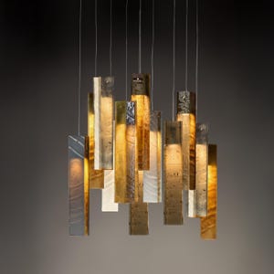 May include: A multi-pendant chandelier featuring numerous rectangular glass panels suspended at varying heights. The panels display a mix of amber, gold, and translucent white tones with textured, organic surfaces that diffuse warm light downward.