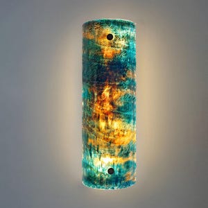 May include: A cylindrical wall sconce featuring a marbled pattern of deep teal, blue, and warm amber tones. The fixture has a translucent, textured surface with two dark circular mounting points positioned at the top and bottom center.