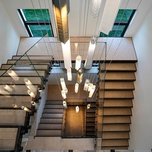 May include: A multi-level staircase with light brown carpeted treads and glass railings, featuring a central chandelier with numerous vertical, rectangular, translucent light fixtures suspended at varying heights from the ceiling above.