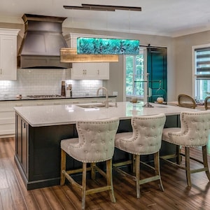 Puede incluir: Una isla de cocina con encimera blanca y base negra, con tres taburetes de bar beige capitoné con tachuelas. Sobre la isla cuelgan dos lámparas colgantes rectangulares, una con acabado dorado texturizado y otra con diseño de vidrio azul verdoso.