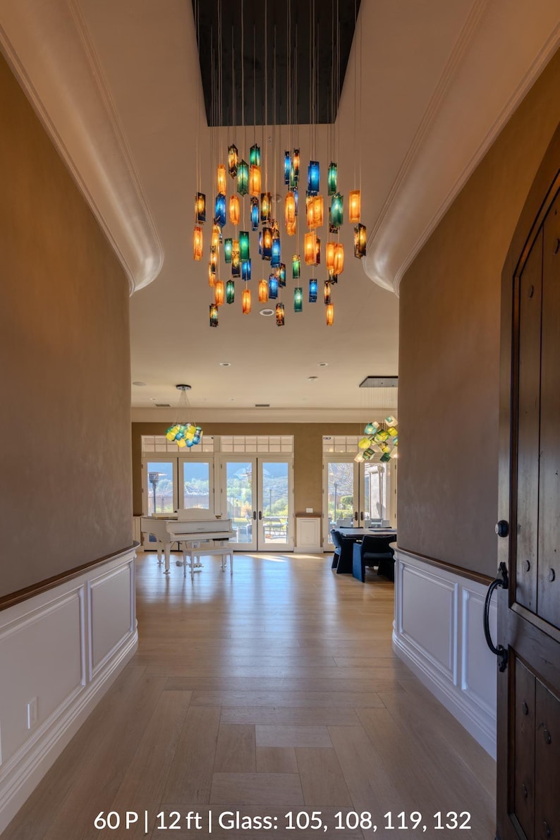 a spacious and well-lit room with a high ceiling, featuring a large chandelier with colorful glass shades hanging from the center. The room has a wooden floor and white walls, creating a warm and inviting atmosphere.