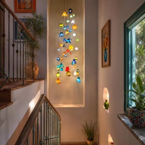 May include: A multi-colored glass pendant light installation featuring numerous small, translucent, bell-shaped and spherical glass elements in shades of blue, yellow, red, green, and white, suspended at varying heights within a recessed wall niche.