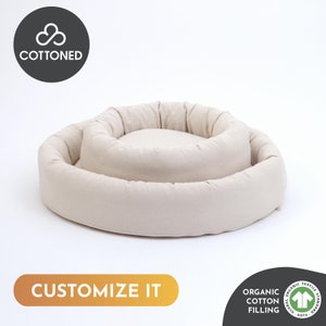 May include: A beige, circular pet bed with a raised outer rim and a lower, central cushion. The bed is made of a soft, textured fabric. The text "CUSTOMIZE IT" is displayed on the bottom left. The text "COTTENED" is displayed on the top left.