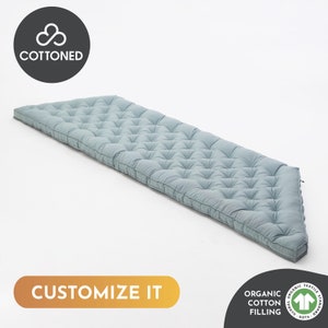 Puede incluir: Un cojín largo y rectangular de color azul claro con mechones. El cojín tiene un patrón de diamantes y está hecho de algodón orgánico. El texto "CUSTOMIZE IT" se muestra en un rectángulo dorado. La imagen también incluye el texto "COTTENED" y "ORGANIC COTTON FILLING".