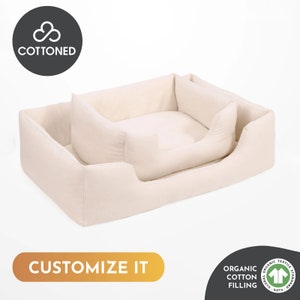 May include: A beige dog bed with a rectangular outer frame and a smaller, cushioned inner bed. The bed has a soft, inviting appearance. The text "CUSTOMIZE IT" is displayed on the bottom. The bed is filled with organic cotton.