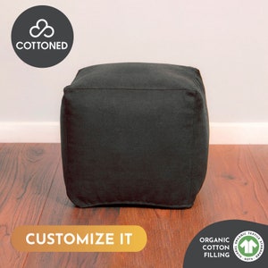 May include: A black square ottoman with a stitched top seam. The ottoman is made of a solid black fabric and sits on a hardwood floor. The text "CUSTOMIZE IT" is displayed on the bottom of the image. The ottoman is filled with organic cotton.