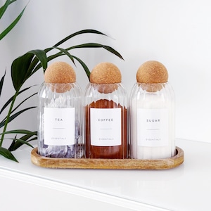 May include: Three glass storage jars with cork lids, labelled "Tea", "Coffee", and "Sugar", are arranged on a wooden tray. The jars contain tea leaves, coffee, and sugar. The word "Essentials" is below each label.